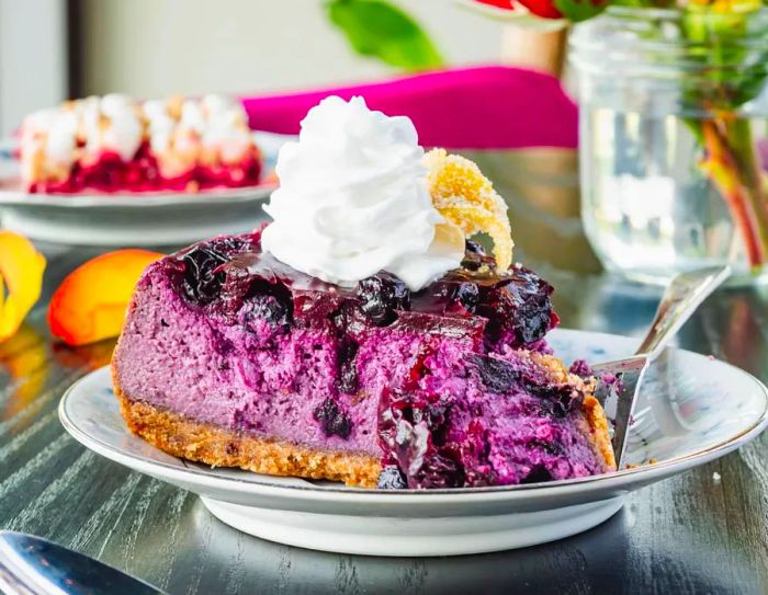 A spoon is poised above a slice of vibrant blue cheesecake, adorned with whipped cream, resting on a table alongside an assortment of other desserts.