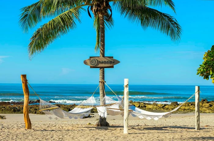 Hammocks draped between frames on a beach beside a wooden sign resembling a surfboard, proudly displaying the name 'Nantipa' on a palm tree.