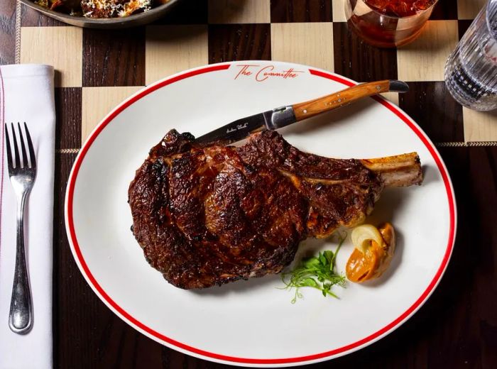 A sizable bone-in steak on an oval platter accompanied by a small scoop of sauce, set on a checkerboard table.