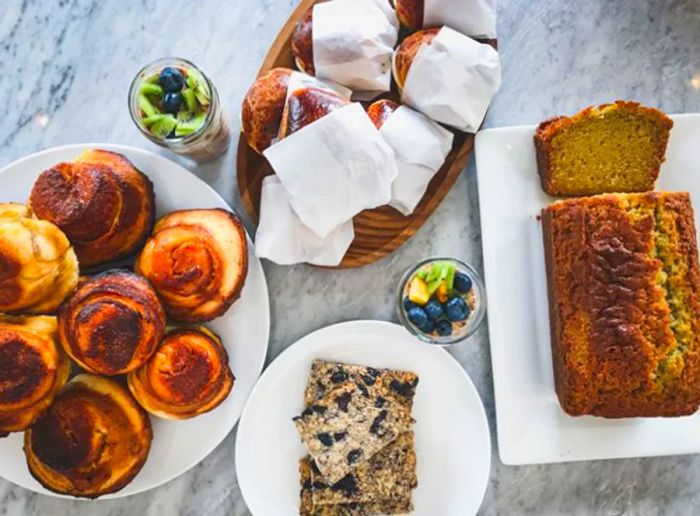 Aerial view of a marble table filled with an assortment of rolls, a loaf of bread, sandwiches, and sweet pastries.