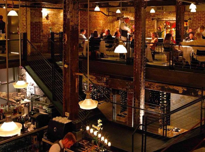 An interior showcasing red brick walls, a two-level layout with large steel beams, metal stairs and railings, tables illuminated by pendant lights, and an open kitchen on the main floor.