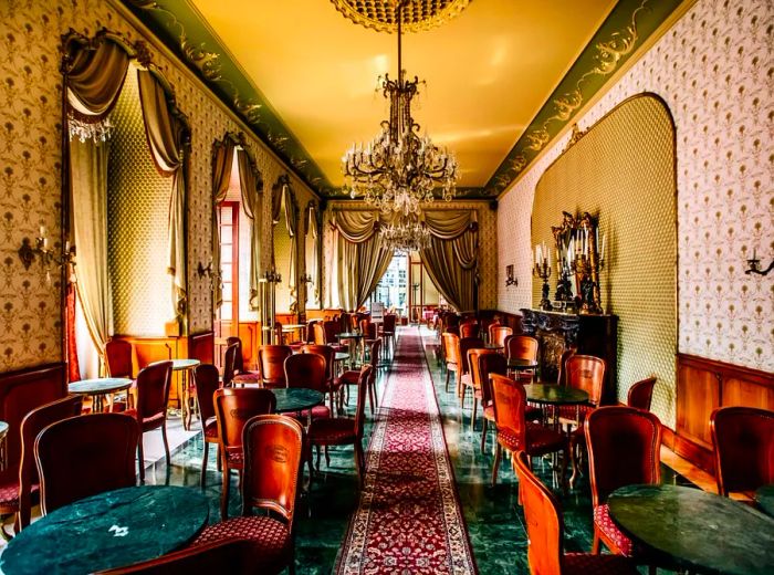 An opulent restaurant interior featuring a long hallway adorned with chandeliers, a textured rug running along the green marble floors, elegant curved-back wooden chairs, and thick drapes framing sunlit windows.