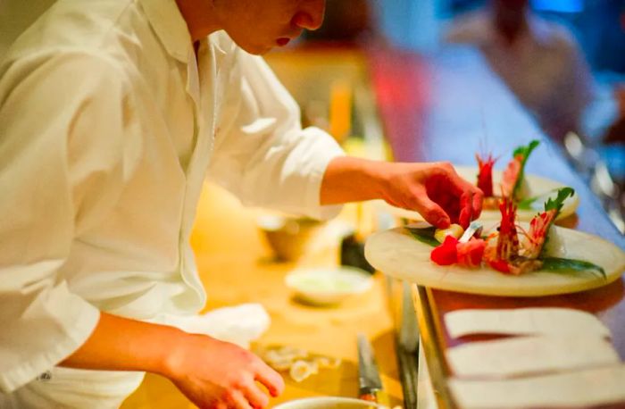 A sushi chef leans over the counter, arranging items on a plate of sashimi.