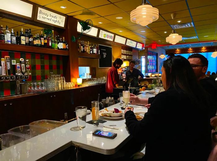 A nostalgic diner interior where patrons gather at the counter relishing food and milkshakes, while staff bustle behind the bar amid patterned wallpaper, midcentury light fixtures, and vintage advertisements.