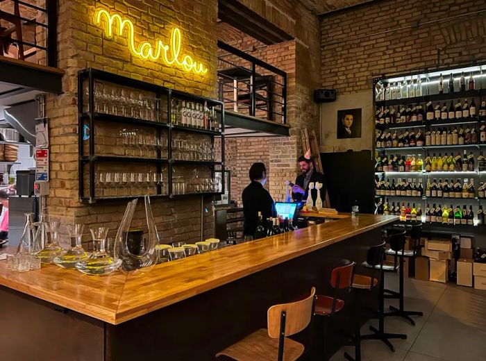 A bartender operates at a computer station behind the bar, where a prominent yellow neon sign proudly displays the name Marlou.