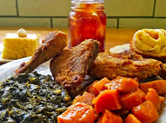 A paper plate filled with pieces of fried chicken alongside heaps of collard greens and yams, complemented by a Mason jar of iced tea and a slice of cornbread in the background.