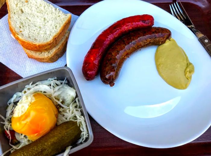 A plate featuring two sausages accompanied by mustard, along with a bowl of pickled vegetables and slices of bread.