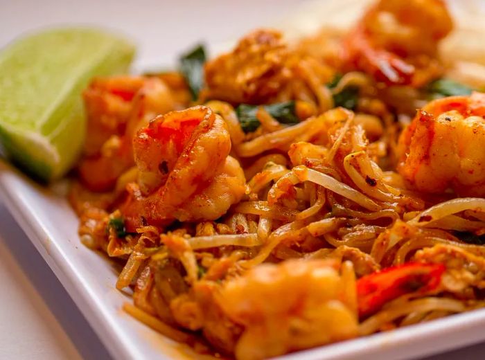 A bowl of pad thai featuring large grilled shrimp and a lime wedge set against a white backdrop.