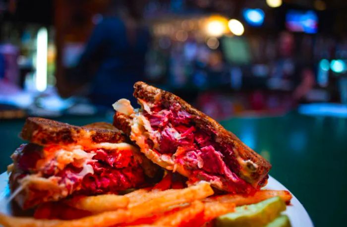 A Reuben sandwich cut in half, resting on a plate alongside a heap of fries, with a softly blurred bar scene in the background.