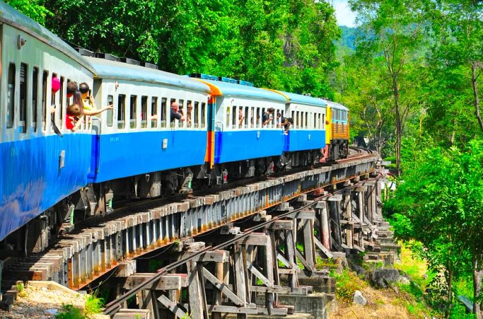 Tourists enjoy the scenic view as they lean out of a train window while crossing a bridge in the Thai countryside.