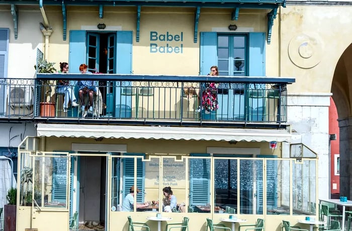 Patrons are seated at outdoor tables on a street-level patio and a balcony patio. The name Babel Babel is displayed on the side of the building.