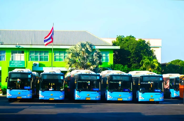 A lineup of blue buses stationed outside a building, adorned with the red, white, and blue striped Thai flag overhead.