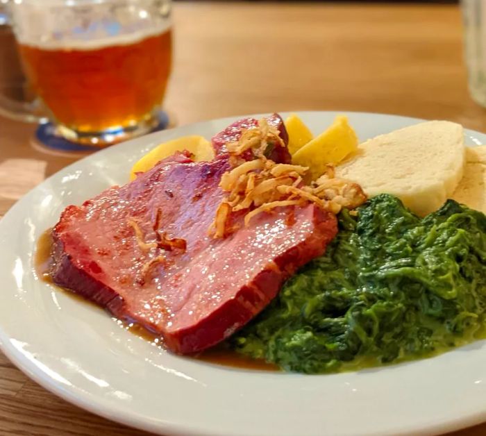 A serving of pork shoulder resting on a bed of cooked spinach, accompanied by slices of bread and a beer.