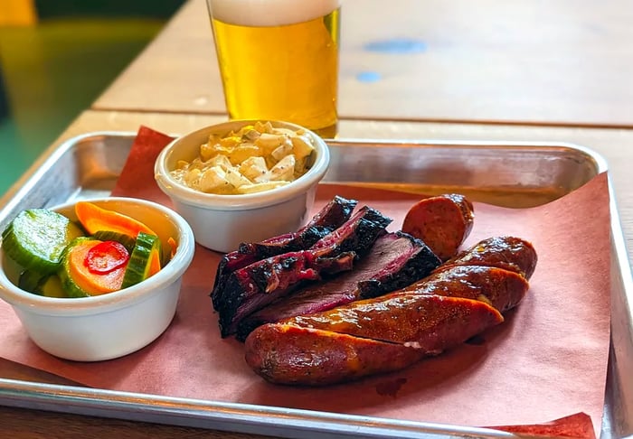 A tray piled with sausages and slices of brisket, accompanied by cups of sides and a glass of beer.