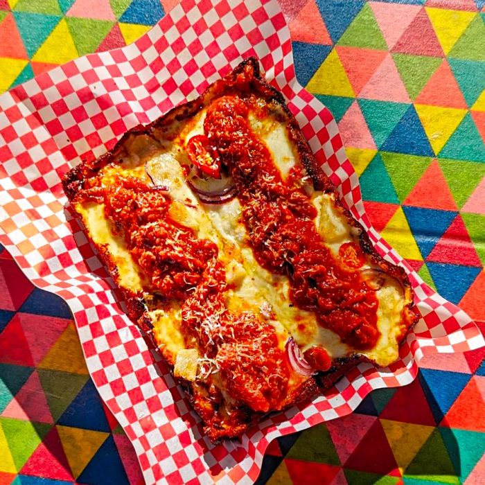 Two elongated strips of pizza, generously slathered with thick tomato sauce, served in a checkered paper-lined boat against a vibrant, rainbow-textured background.