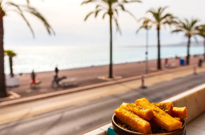 A bowl of panisse sits before a picturesque view of the street, palm trees, and the sparkling sea beyond.