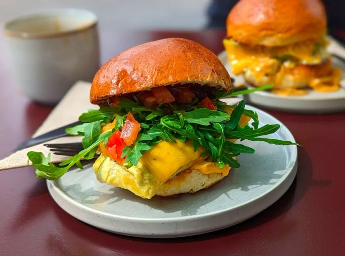 A breakfast sandwich overflowing with arugula, sliced tomato, melted cheese, and a fluffy omelet.