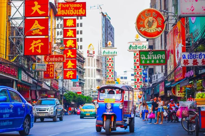 Cars and tuk-tuks navigate a bustling street adorned with vibrant neon signs