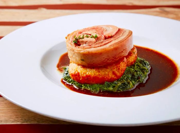 A portion of rolled meat resting atop a fried item, accompanied by a bed of cooked greens.