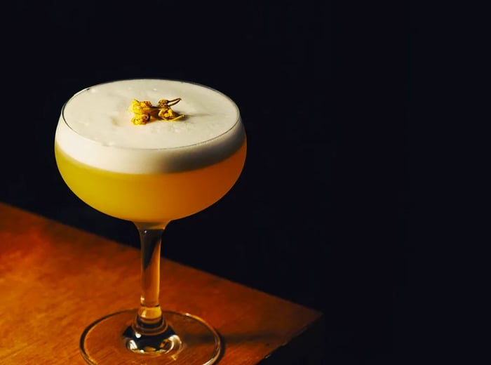 A vibrant yellow cocktail served in a coupe glass, topped with a frothy head and adorned with a small herbal garnish.