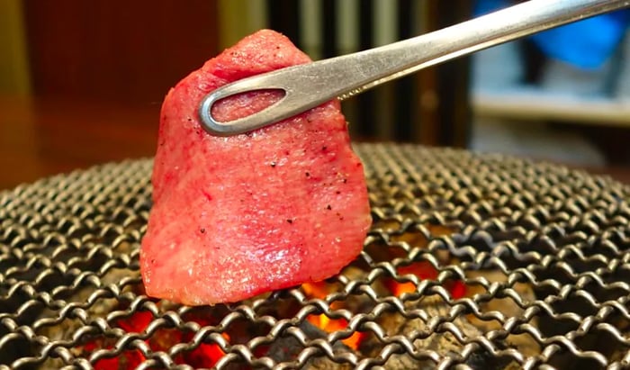 Tongs deftly flip grilled tongue on a charcoal grill.