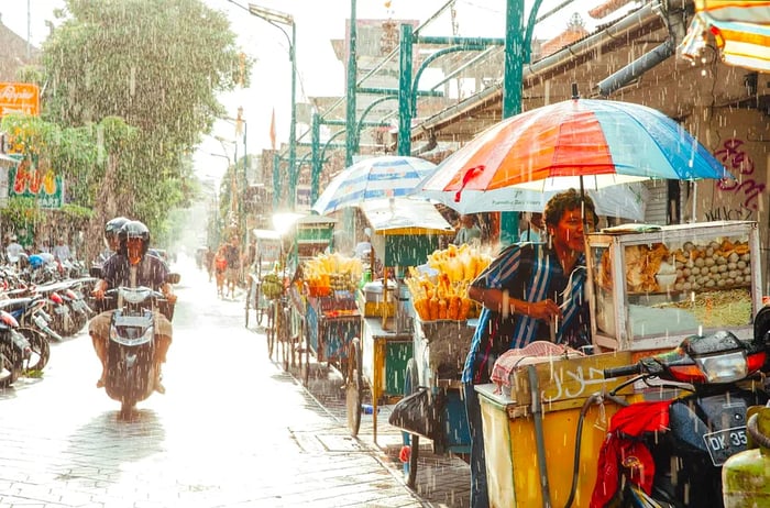 A motorcycle carrying a passenger navigates a street filled with food stalls as rain falls, while the sun peeks through the clouds.