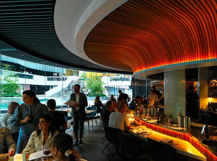 A dining space featuring a wavy, illuminated ceiling and a wall lined with windows. Servers navigate between tables and the bar area where guests are seated.