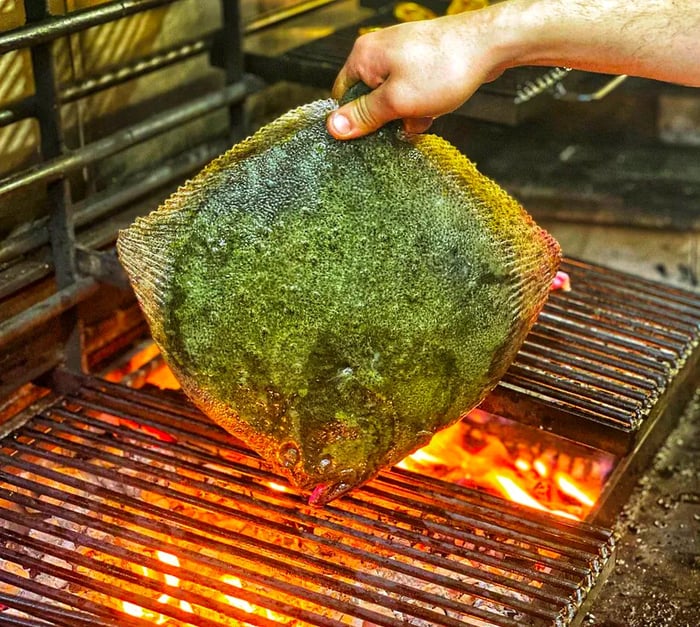 A hand lifts a whole fish above a wood-fired grill.