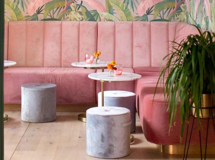 A cozy bar area featuring pink banquettes nestled in the corner, adorned with tropical pink wallpaper. Scattered around are small marble tables and short stools that resemble concrete, complemented by a large plant in one corner.