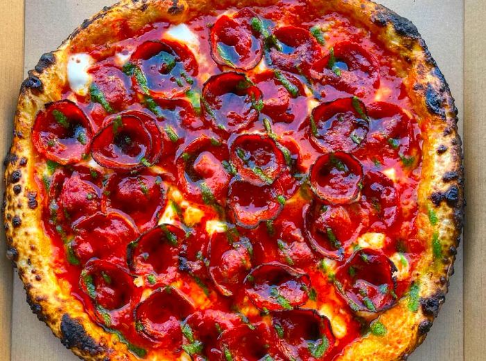 A bird's-eye view of a complete pepperoni pizza adorned with stripes of green sauce and a sheen of oil.