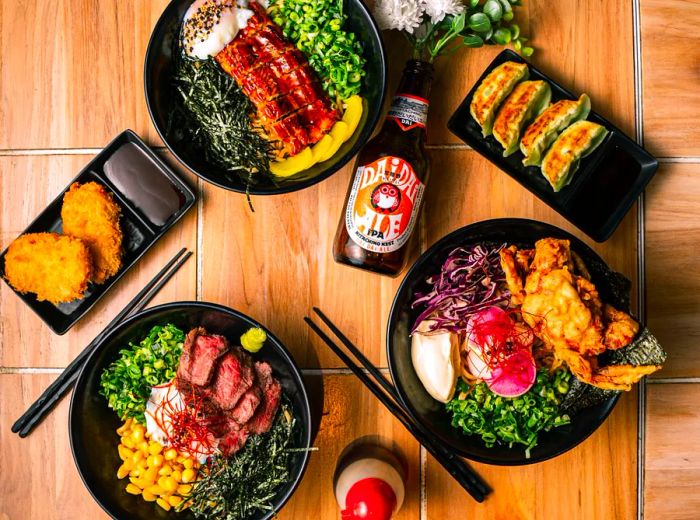 Three bowls of soba noodles adorned with vibrant toppings, accompanied by a variety of dishes and a bottle of rice beer.