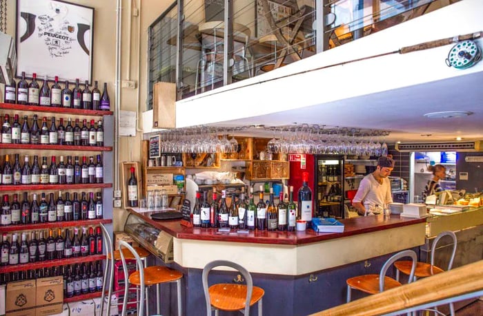 Bartenders behind a bar lined with bottles, adjacent to a large shelf of more bottles, with an upstairs seating area visible above.