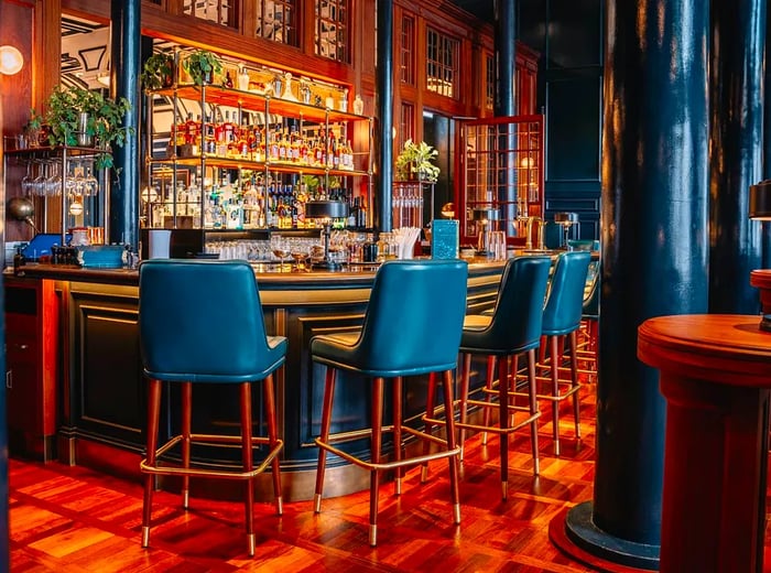 A luxurious bar featuring deep blue leather stools, illuminated shelves brimming with spirits, and rich wooden accents.