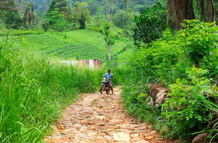 A young local man skillfully rides his motorcycle along a narrow, bumpy road in the small town of Deniyaya, Sri Lanka, with lush greenery surrounding the path.