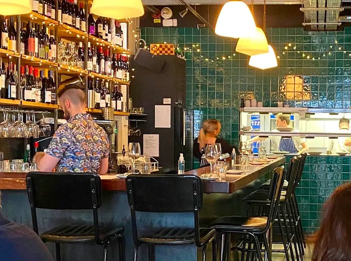 A bar adorned with dark green tiles and pendant lights, where bartenders are busy at work.