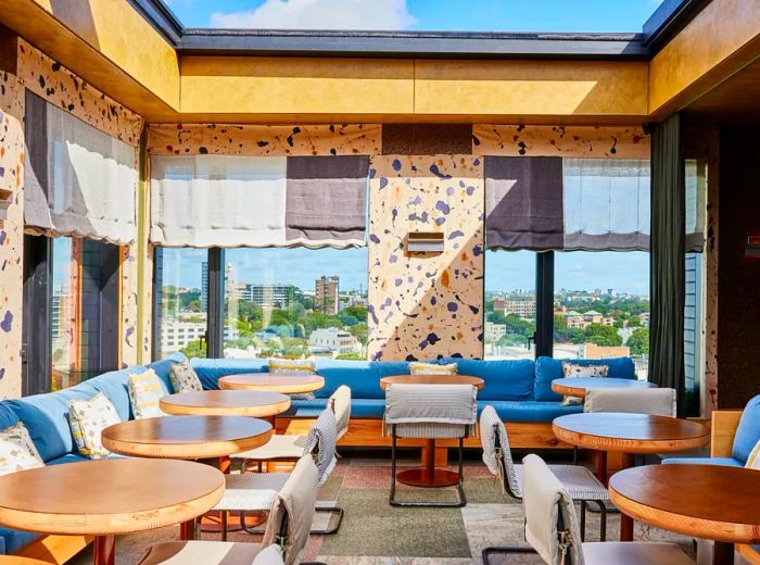 A vibrant rooftop dining space featuring quirky patterned walls and expansive windows that showcase stunning city views, complete with curvy midcentury chairs and cozy banquettes.