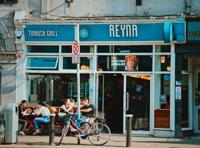 Patrons enjoy their meals at tables outside a restaurant adorned with signs for Reyna and Turkish Grill.