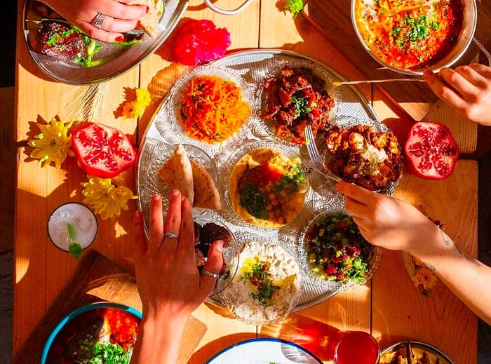 Hands reaching into a variety of vibrant dishes atop a sun-drenched wooden table.