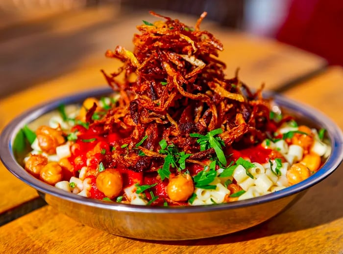 A towering heap of crispy fried onions crowns a metal bowl of koshari, accompanied by browned chickpeas and fresh herbs.