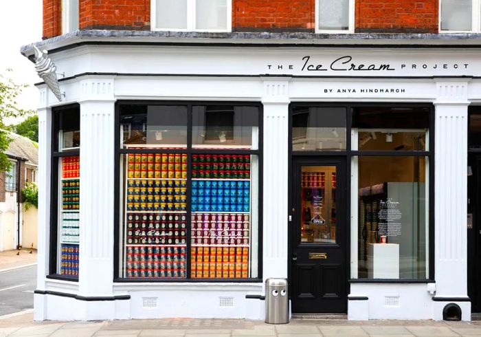 The exterior of a white store features large windows displaying pints of ice cream stacked in tall towers, with The Ice Cream Project sign prominently above the entrance.