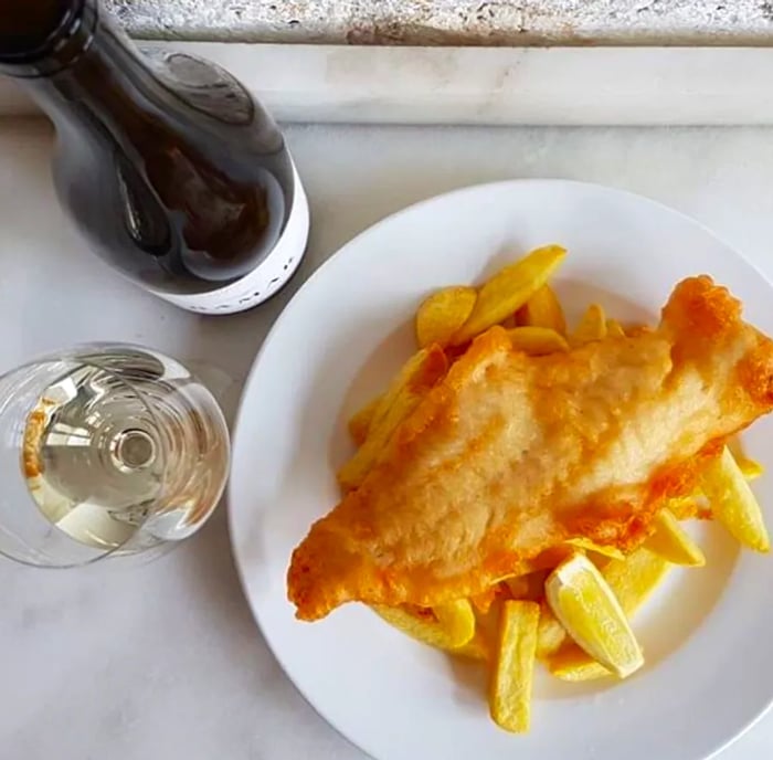 A plate of fried fish served over a bed of fries, accompanied by a glass of white wine and a bottle.