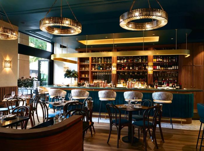 An elegant bar and dining area adorned with grand chandeliers, a golden railing above the bar, mid-century stools, and polished hardwood floors.