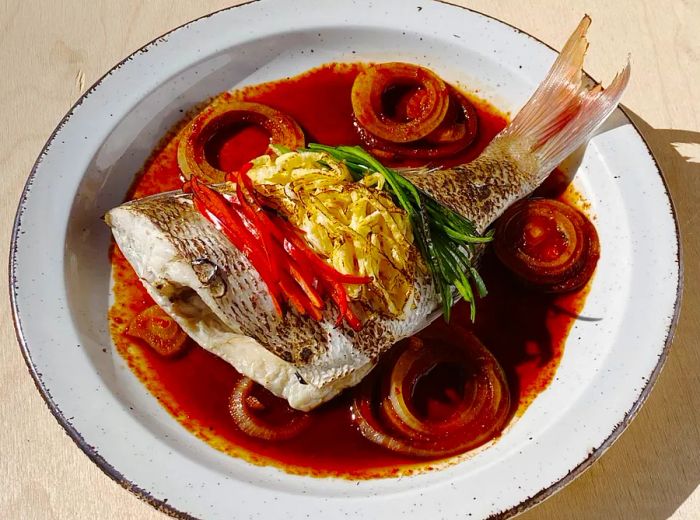 A generous portion of fish with its tail, resting in a vibrant deep red sauce and adorned with slices of cooked vegetables.