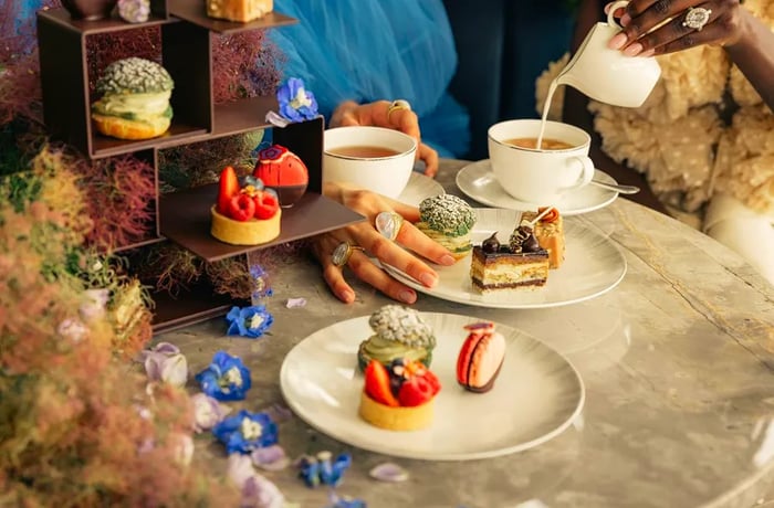 Guests relish beautifully crafted pastries paired with cups of tea on a marble table surrounded by dried foliage and a stack of additional treats.