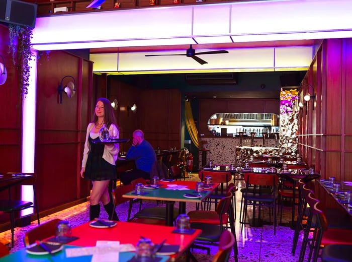 A waiter navigates through a dining area adorned with purple lighting, a patterned floor, and large tubular fixtures.