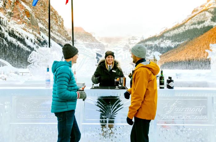 Two patrons stand at an ice bar, attended by a bartender, with flags fluttering in the background and sunlit mountains in the distance.