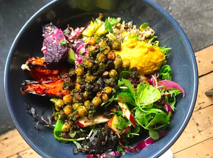 A hand cradles a dark bowl brimming with an assortment of vegetable, chickpea, and herb dishes artfully arranged together.