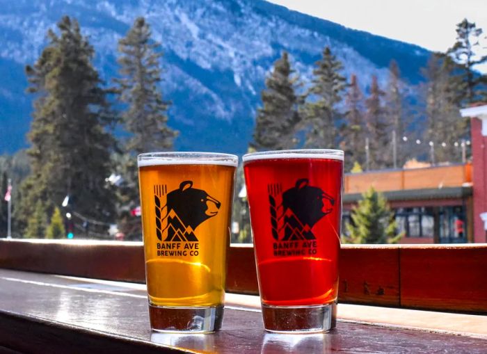 Two glasses of differently colored beer sit on a bar in front of a stunning mountain backdrop.