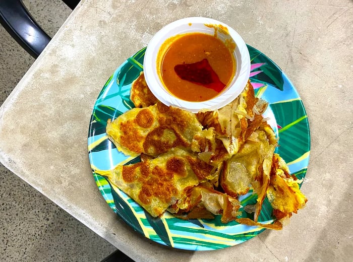 Torn pieces of roti arranged on a plate featuring a tropical leaf design, accompanied by a disposable bowl of dipping sauce.