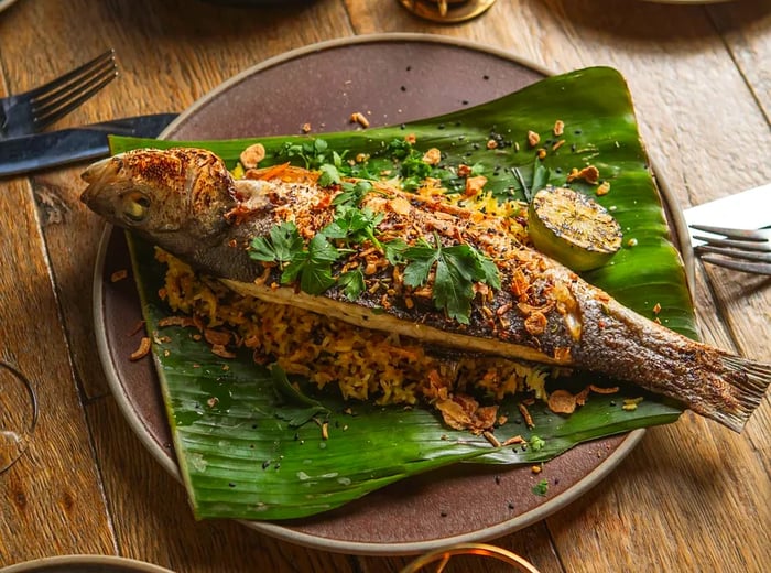 A whole fish garnished with crispy toppings and cilantro, elegantly served on a leaf.